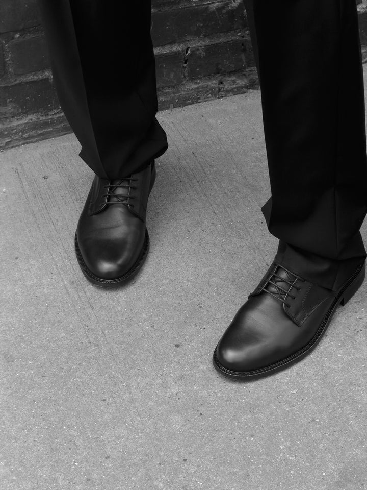 Black Legacy Derby shoes worn with dark trousers beside a brick wall, photographed in black and white.