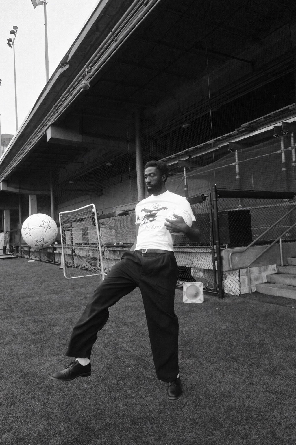 Black-and-white photograph of a man in tailored trousers juggling a football on the pitch