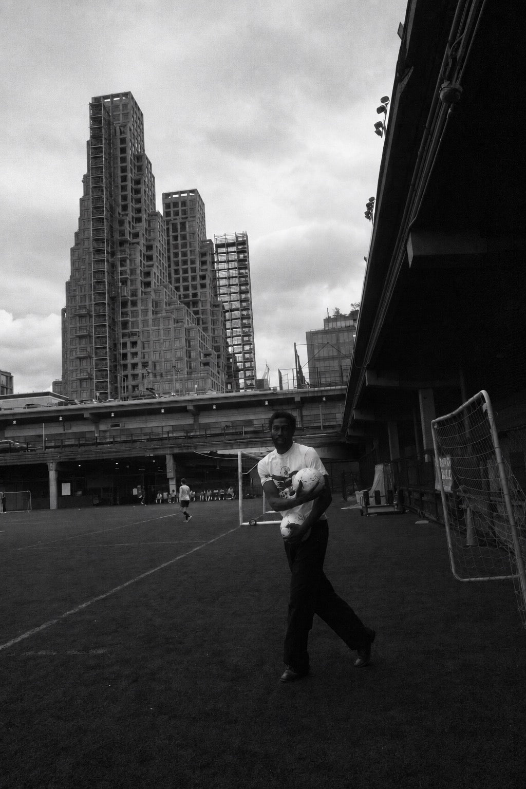 Black-and-white wide view of a man carrying footballs across a pitch beneath a bridge beside tall city buildings