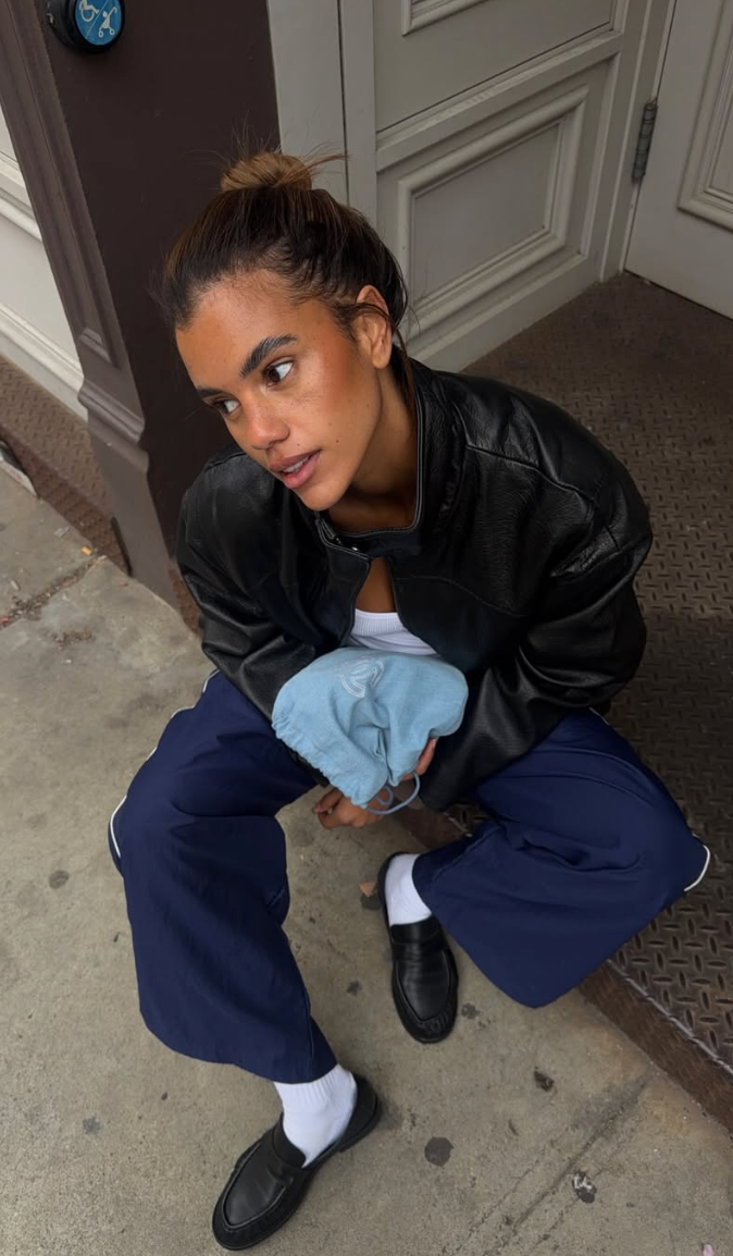 A seated woman in a black leather jacket and navy trousers, wearing black loafers with white socks.