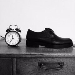 Photographie en noir et blanc d’une chaussure noire à lacets et d’un réveil sur un meuble en bois.