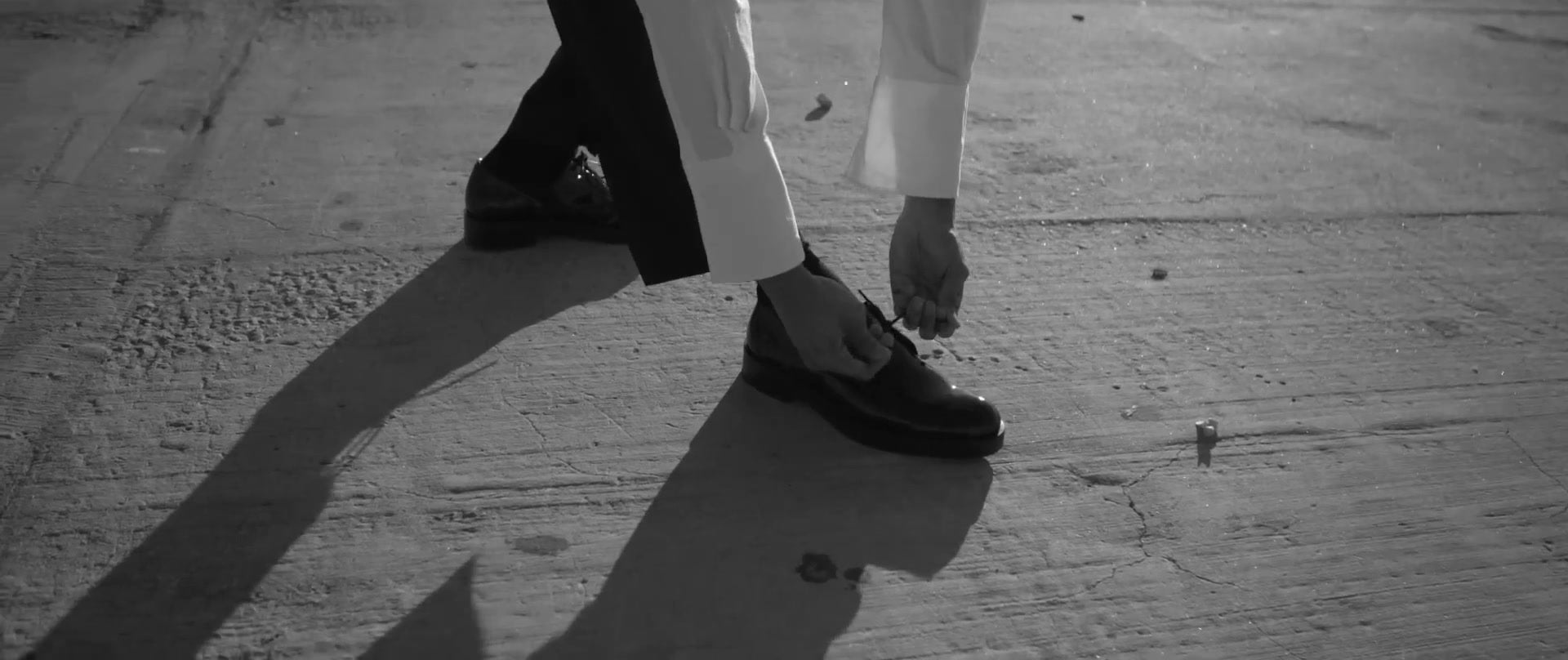 Une personne noue les lacets d’une chaussure noire ; de longues ombres se dessinent sur le béton.