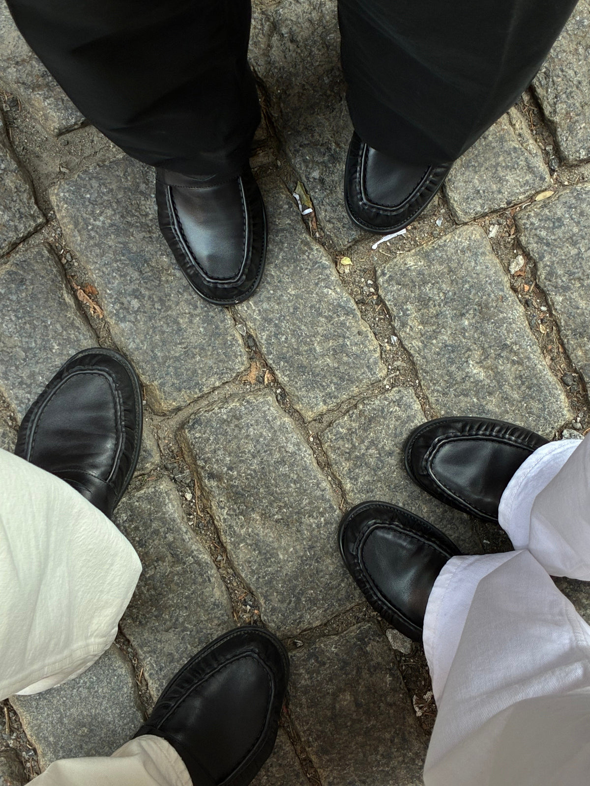 Three pairs of black moccasins worn together on a cobbled pavement.
