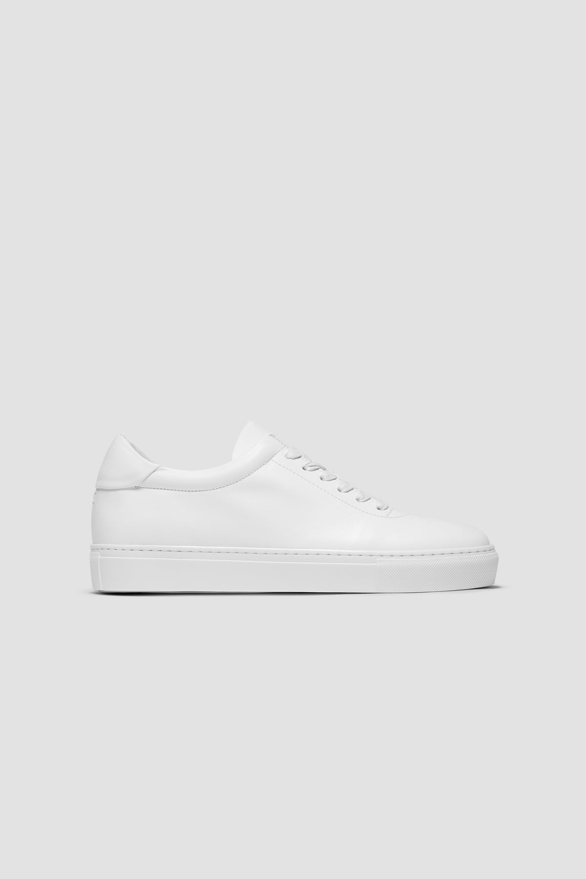 The Proper Sneaker® in white leather, seen in profile against a light-grey studio background.