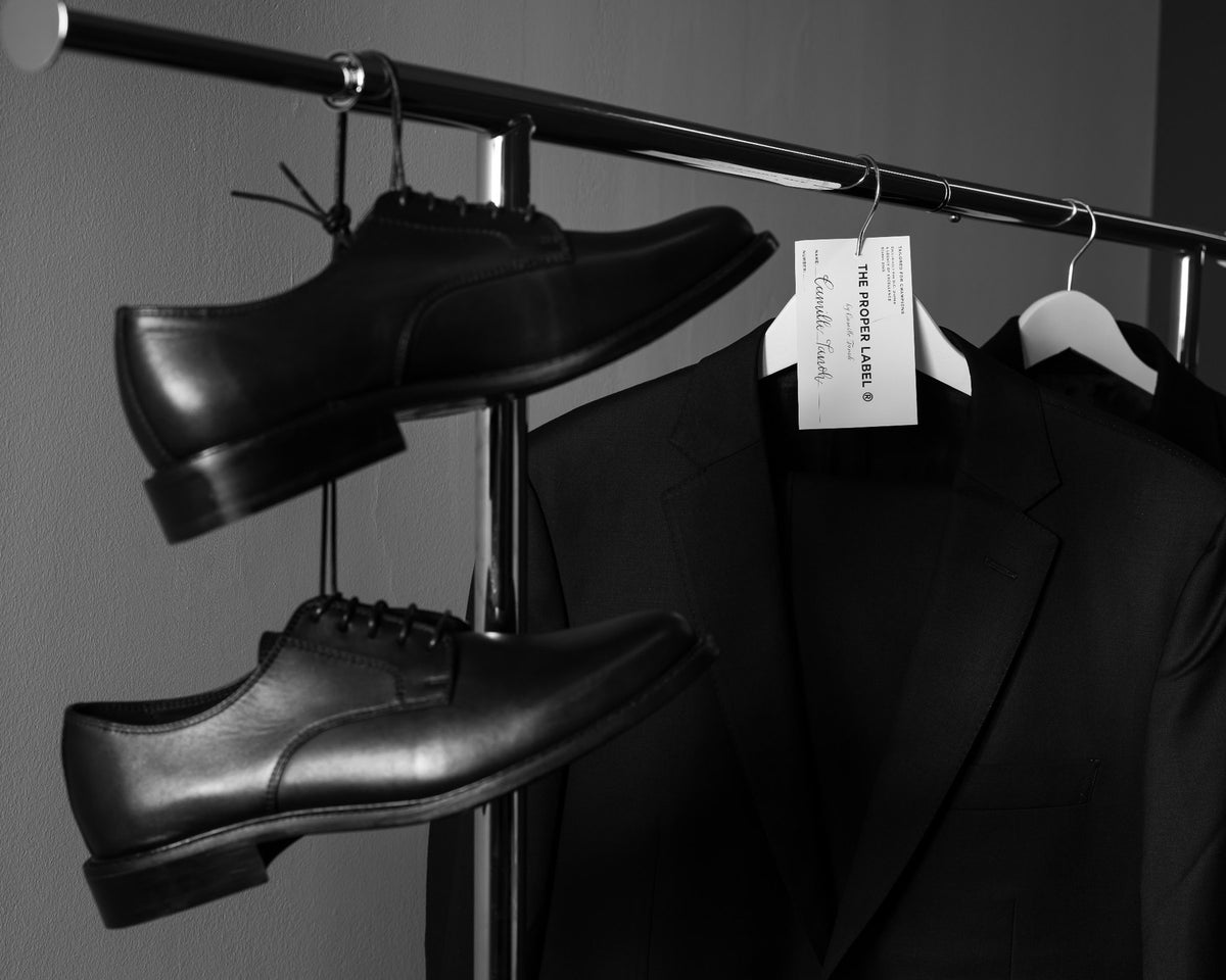 Historical black lace-up shoes suspended beside jackets and a fitting tag. Maison de Sport — Collection 005. Photography: Malike Sidibé, New York.