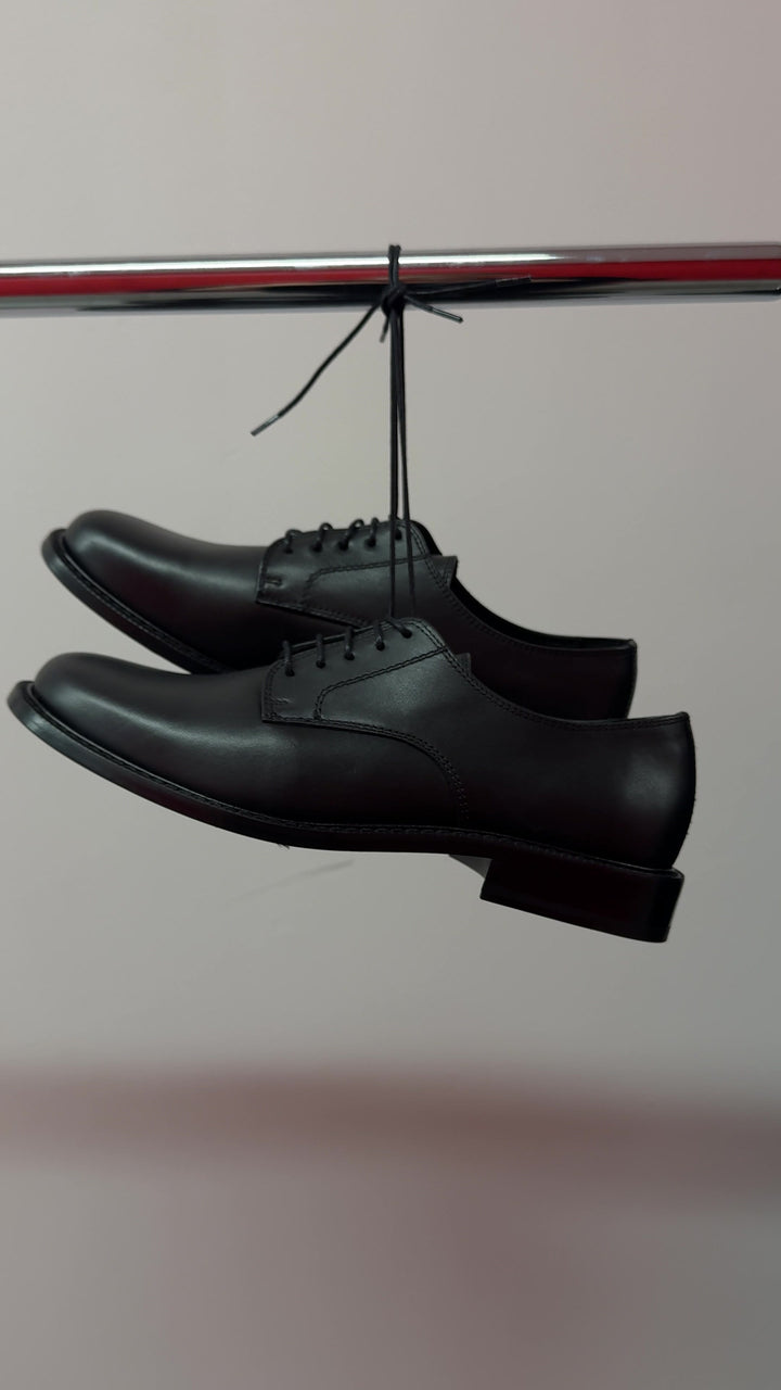 Two black lace-up Derby shoes hanging by their laces from a rail, a still from the supplied film.