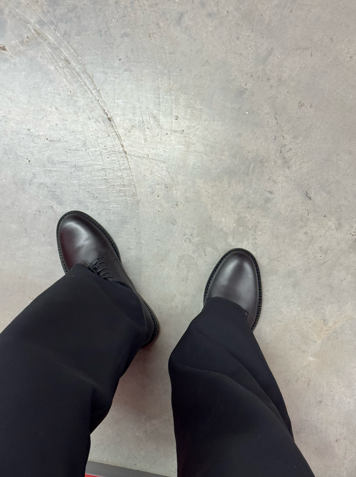 Black lace-up Derby shoes worn with black trousers, photographed from above on a concrete floor.