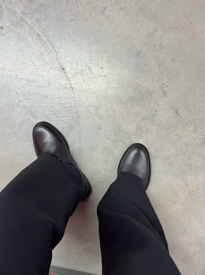 Black lace-up Derby shoes worn with black trousers, photographed from above on a concrete floor.