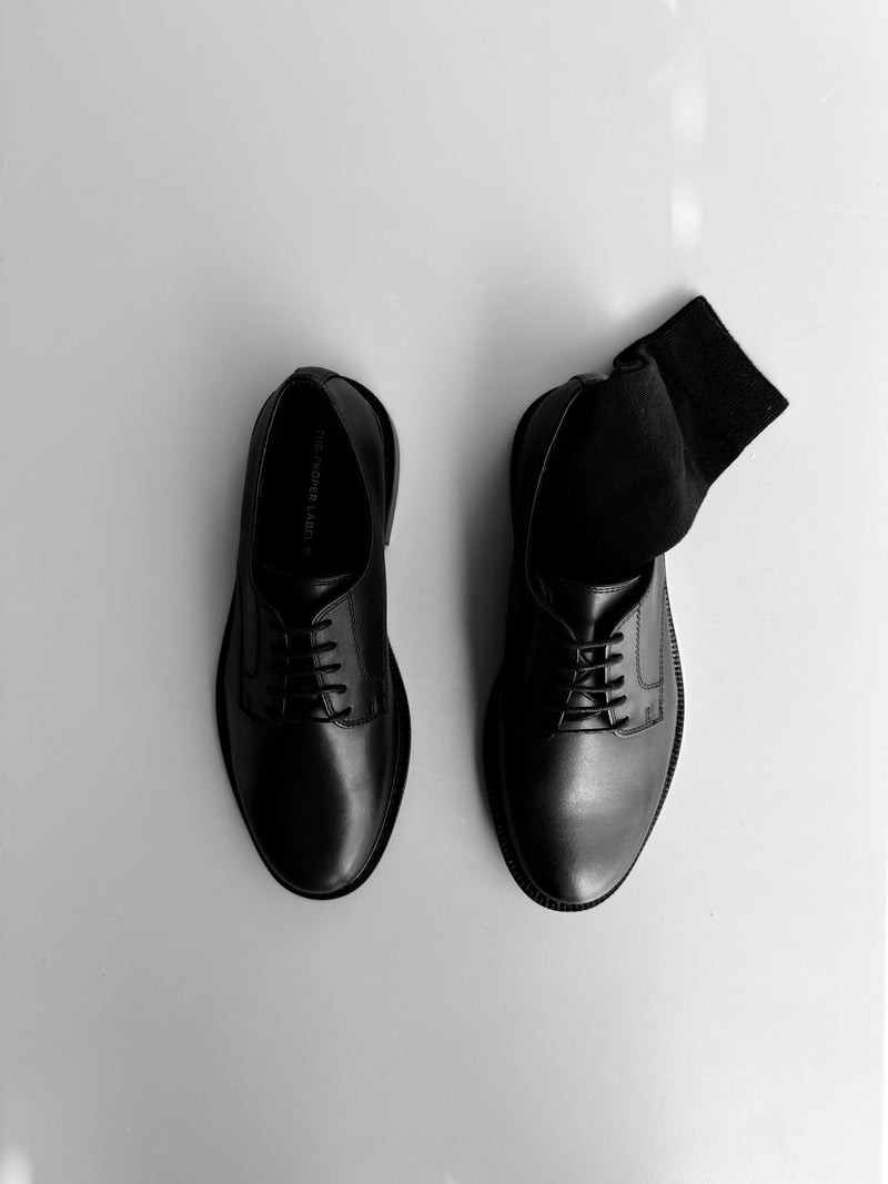 The Legacy Derby in black leather, a pair photographed from above on a pale surface.