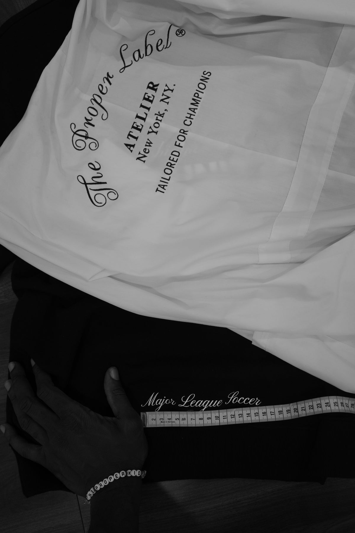 A hand and measuring tape at work on a Major League Soccer garment beside The Proper Label atelier shirt