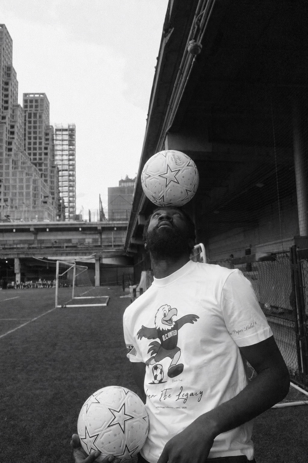 Black-and-white photograph of a man balancing a football on his head while holding a second ball on an urban pitch