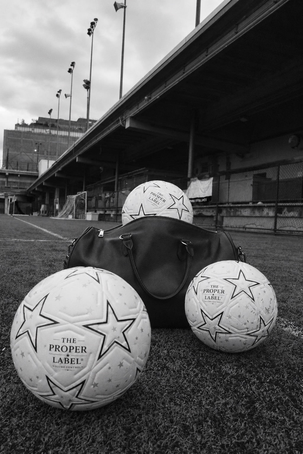 Three The Proper Label footballs and a black leather travel bag on an urban football pitch, in black and white.
