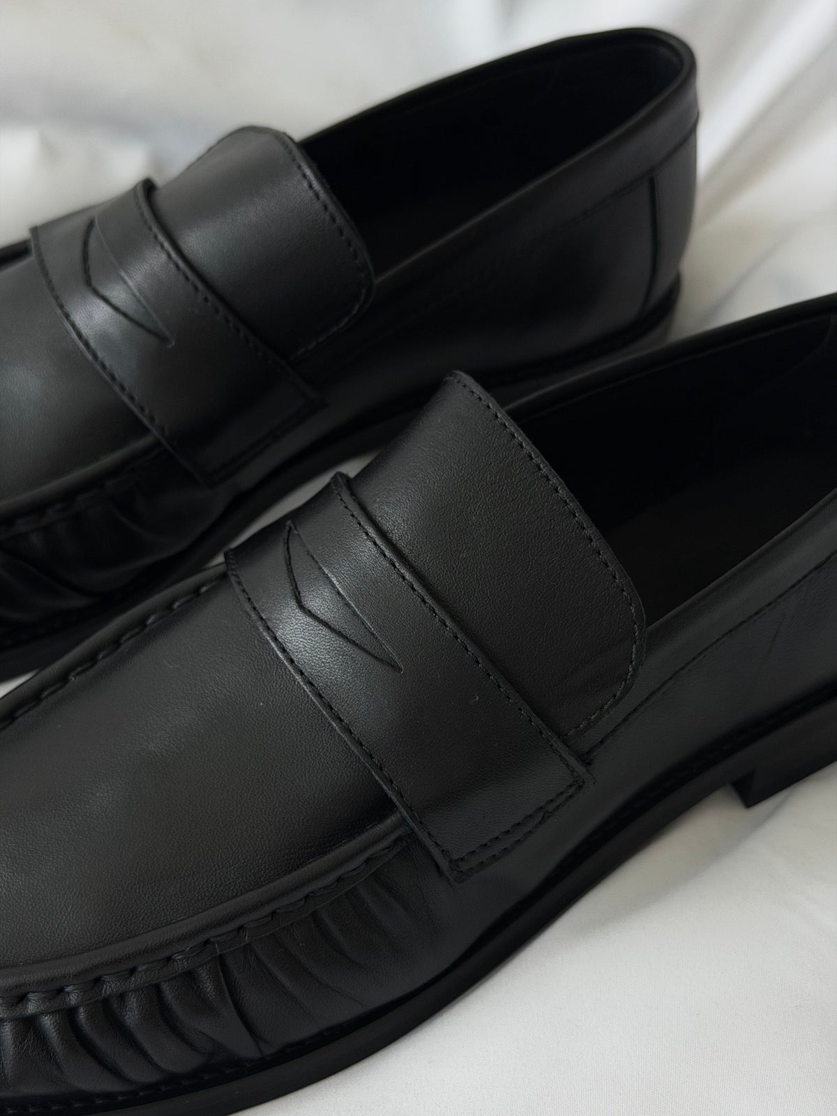 Close-up of black Proper Moccasins on white fabric, showing the leather and signature strap.