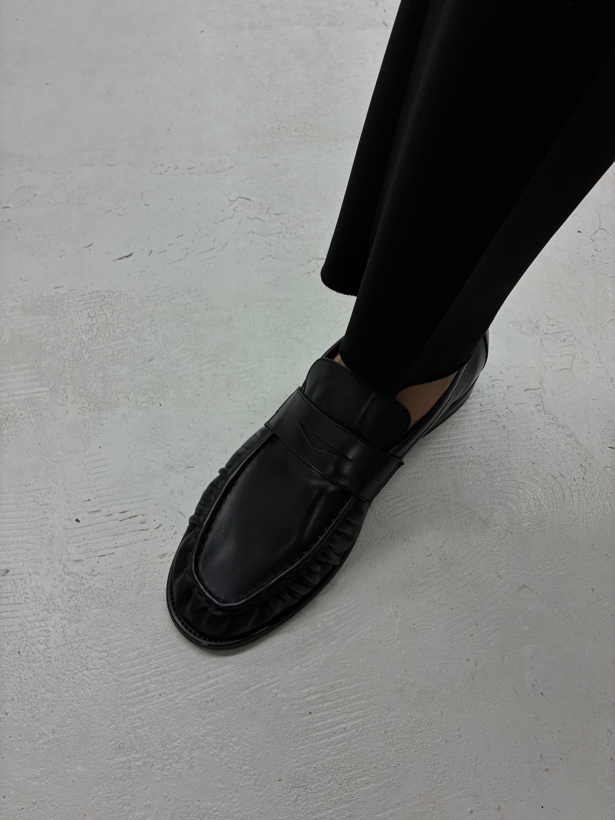 Black Proper Moccasin worn with dark trousers on a pale floor.