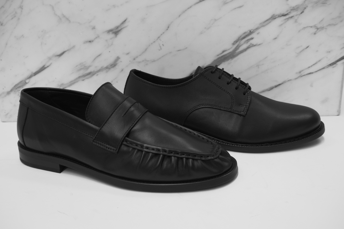 The Legacy Derby and The Proper Moccasin in black leather against veined marble.