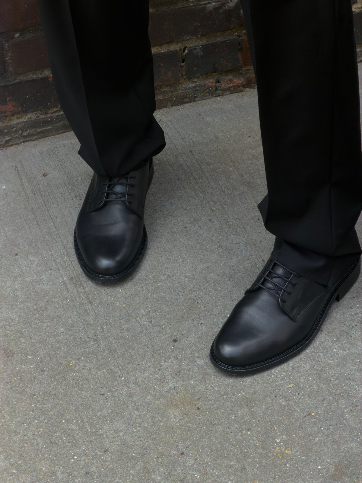 Black leather Derby shoes worn with dark trousers, photographed on a concrete pavement.