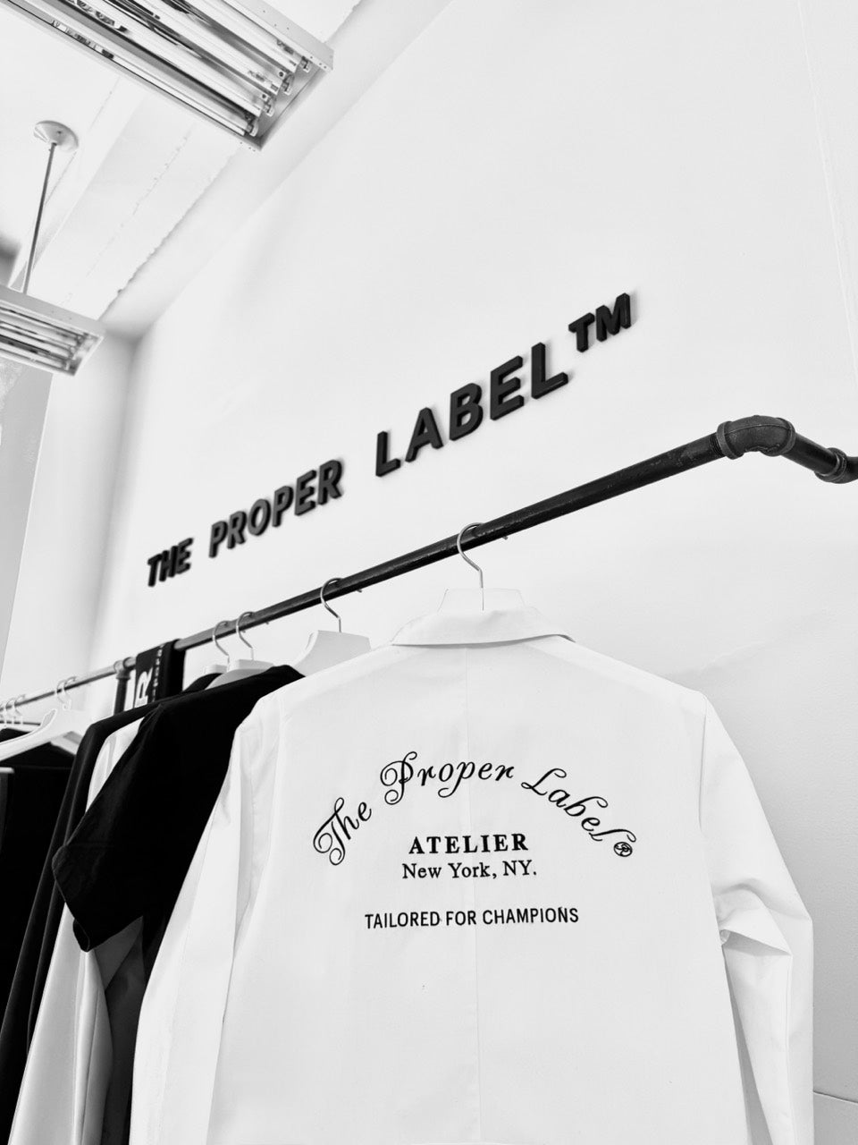 The Proper Label atelier: a white Tailored for Champions shirt on a rail beneath the Maison signature, in black and white.