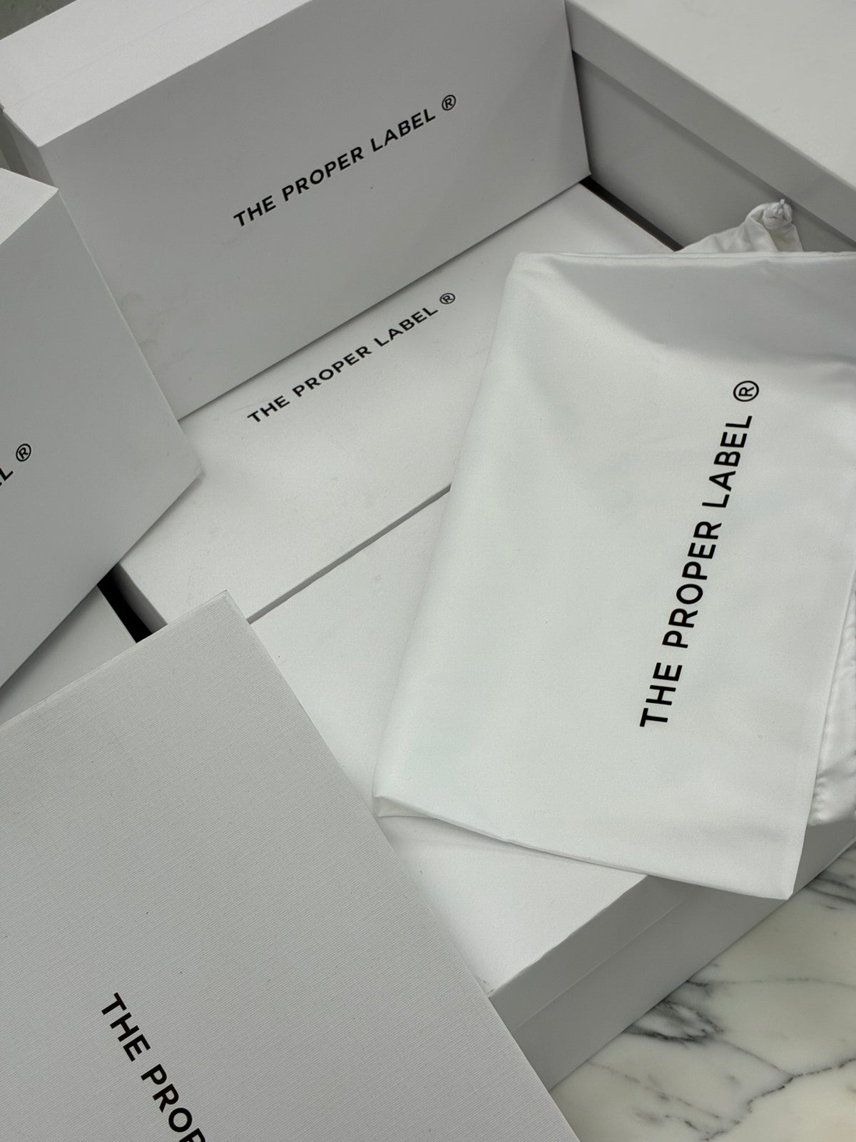 White Proper Label boxes and branded dust bags on marble.