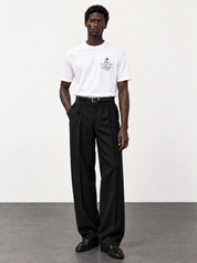 Maison de Sport white T-shirt with black pleated trousers and moccasins, photographed against an off-white studio wall and dark grey floor.
