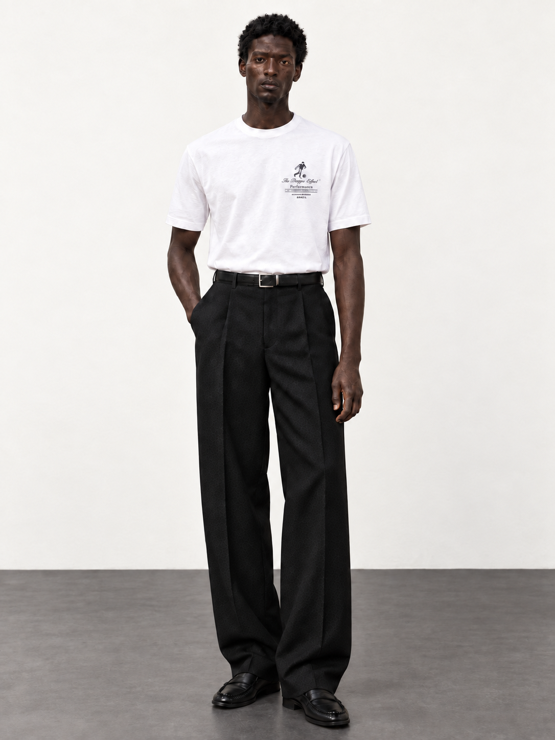 Maison de Sport white T-shirt with black pleated trousers and moccasins, photographed against an off-white studio wall and dark grey floor.