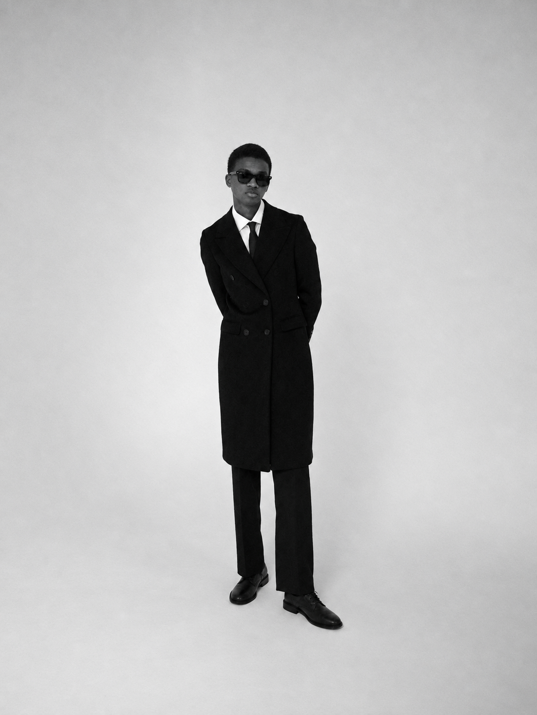 Full-length black-and-white portrait in a black double-breasted overcoat, shirt, tie and lace-up shoes.