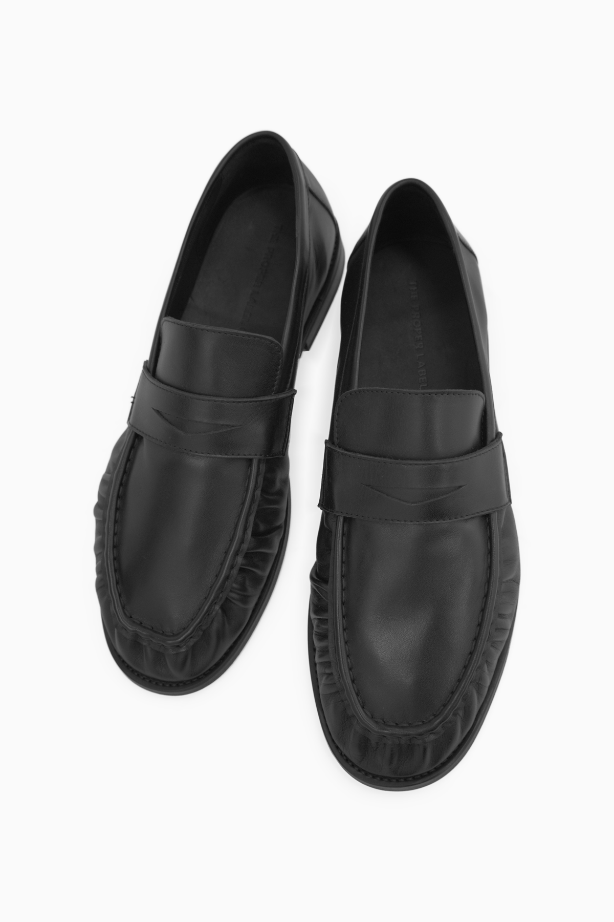 The Proper Moccasin in Obsidian Black, pair viewed from a high angle, with leather saddles and gathered apron stitching visible.