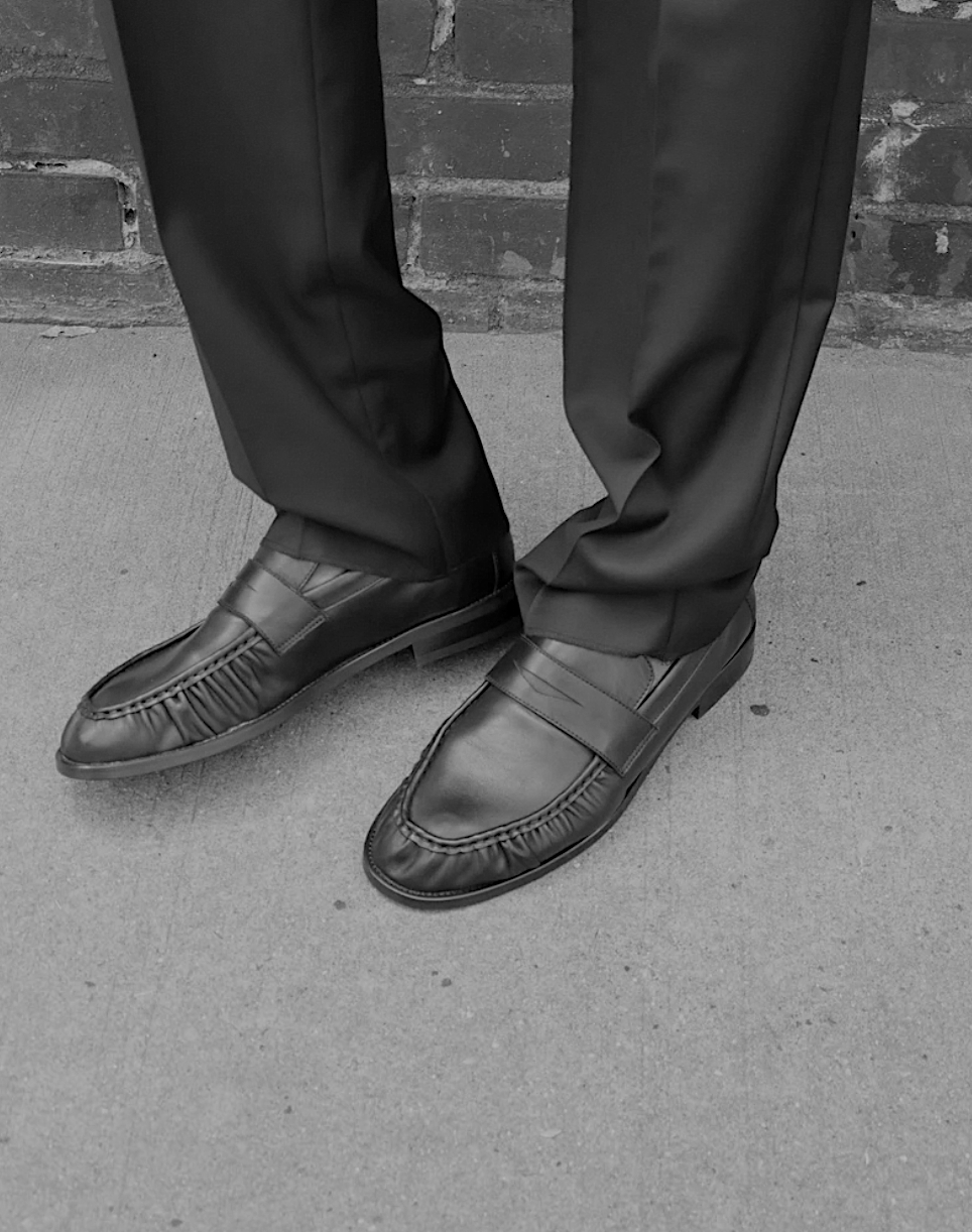 The Proper Moccasin worn with black tailored trousers against a brick wall.