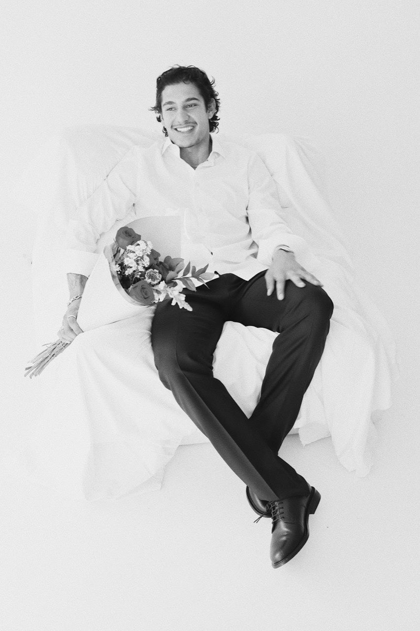Gabriel Robles seated with flowers, wearing a white shirt, black trousers and black lace-up shoes.