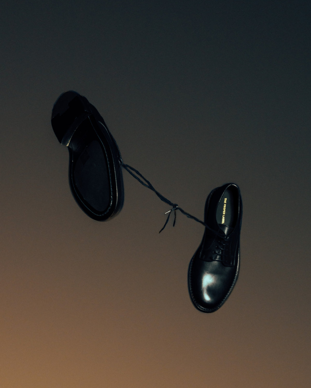 A pair of black leather Derby shoes suspended by their tied laces against a dark background.