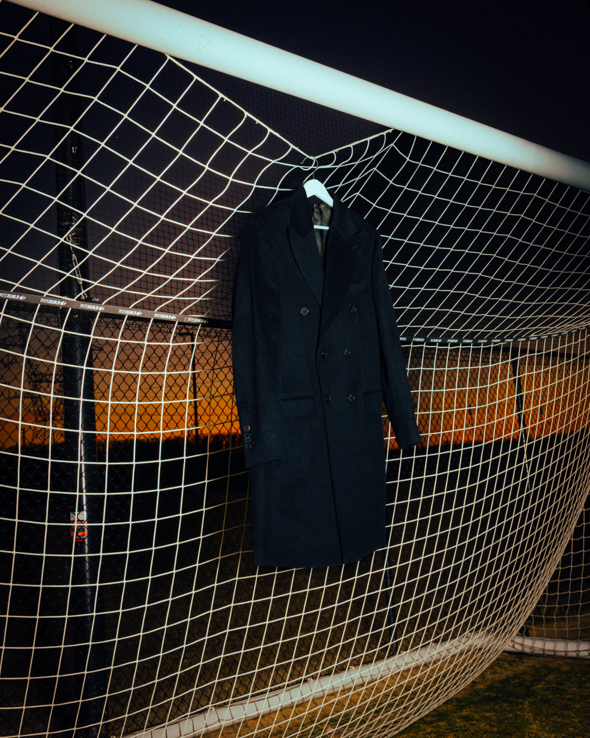 A dark double-breasted overcoat hanging from a white football goal net at night.