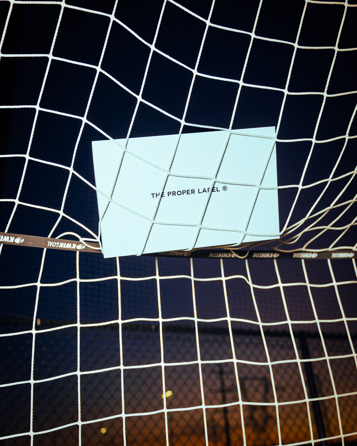 A white box bearing The Proper Label wordmark held in the mesh of a football goal net.