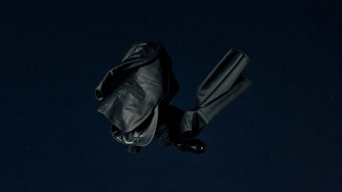 Black tailored jacket, trousers and a derby shoe arranged on a dark background.