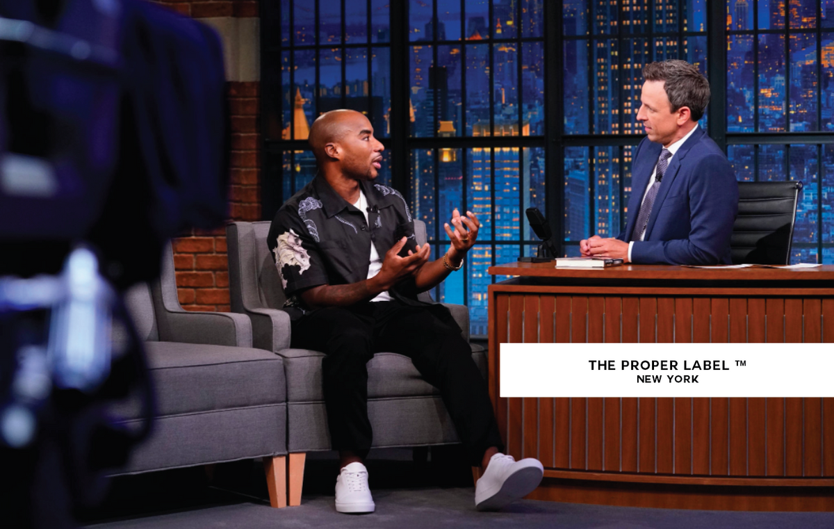 Two people in a television studio; the seated guest wears The Proper Sneaker®. The Proper Label New York branding remains visible.