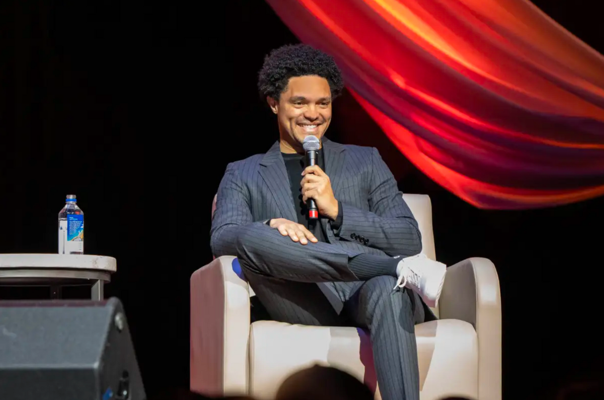 Trevor Noah seated on stage with a microphone, wearing a suit and white Proper Sneakers.