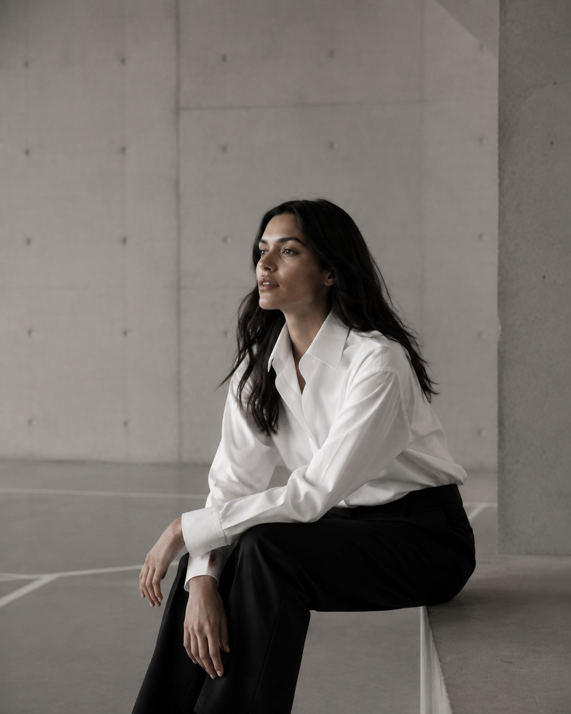 A woman in white cotton shirting seated in a softly lit concrete sports hall.