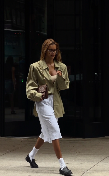 A woman walking in an olive overshirt and light skirt, wearing black shoes with white socks.