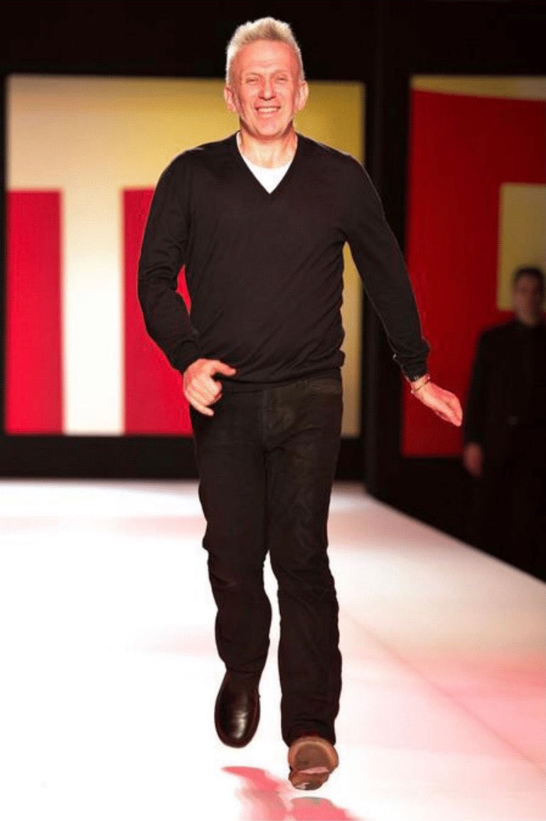 Jean Paul Gaultier s’avançant sur un podium, vêtu de sombre, les deux chaussures visibles.