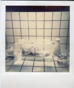 Polaroid of a sneaker among glass vessels on a tiled surface.