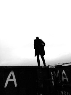 Photographie en noir et blanc d’une personne en silhouette au bord d’un toit, au-dessus d’un mur portant des lettres et des chiffres peints.